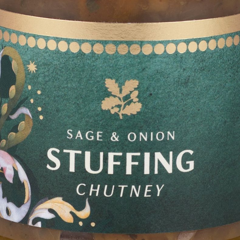 Close-up of a jar of sage and onion stuffing chutney, showing a dark green label with white product lettering, gold decorative accents and festive botanical artwork, with chunky chutney visible through the clear glass jar above and below the label.