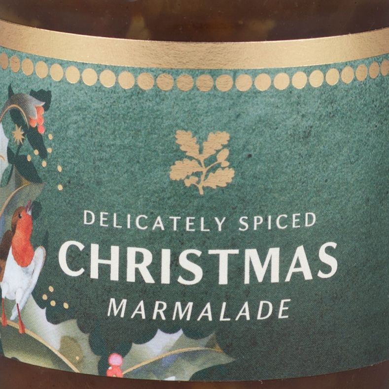 Close-up of a jar of delicately spiced Christmas marmalade, showing a dark green label with white product lettering, gold decorative accents and festive robin and holly artwork, with marmalade visible through the clear glass jar above and below the label.