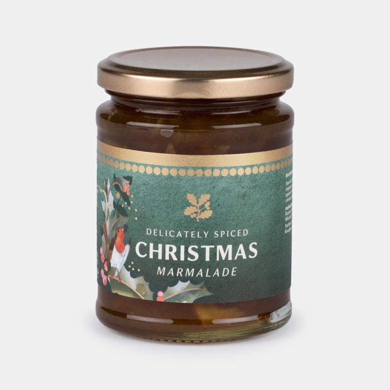 Jar of delicately spiced Christmas marmalade, featuring a clear glass jar with a gold lid and a dark green label decorated with festive robin, holly and botanical artwork, showing marmalade inside and isolated on a white background.