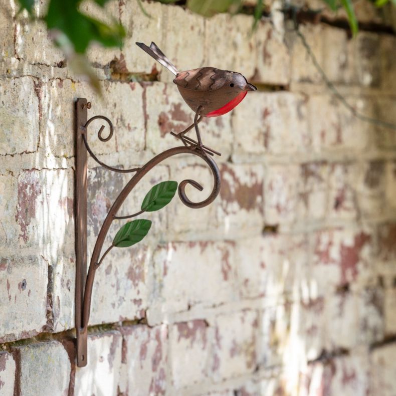 Metal robin hanging basket bracket featuring an antique copper-effect finish, decorative scrollwork support arm, green metal leaf accents, and a sculpted robin with a red breast and engraved feather detailing perched on top, shown mounted against a rustic