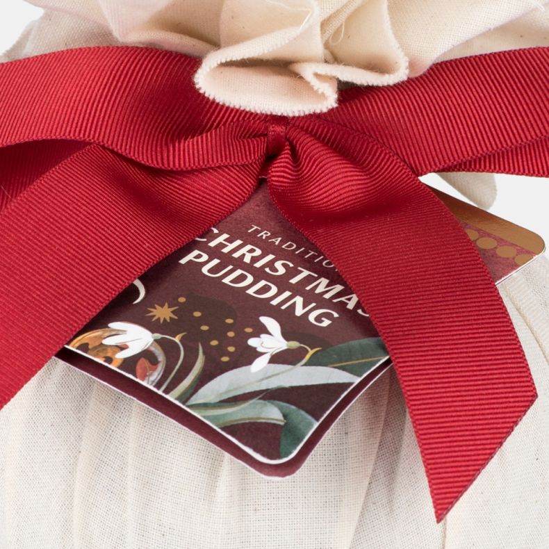 Close-up of a traditional Christmas pudding wrapped in natural cloth, featuring a red ribbon bow and festive gift tag with seasonal botanical illustrations.