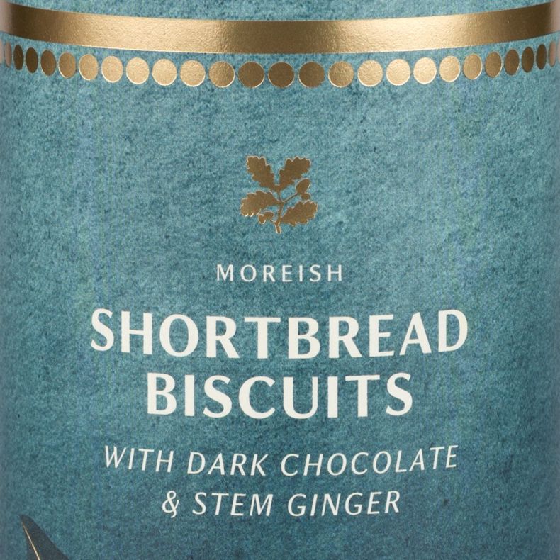 Close-up of a decorative teal biscuit tin label displaying the text “Shortbread Biscuits with Dark Chocolate & Stem Ginger” with gold detailing and festive botanical illustrations.