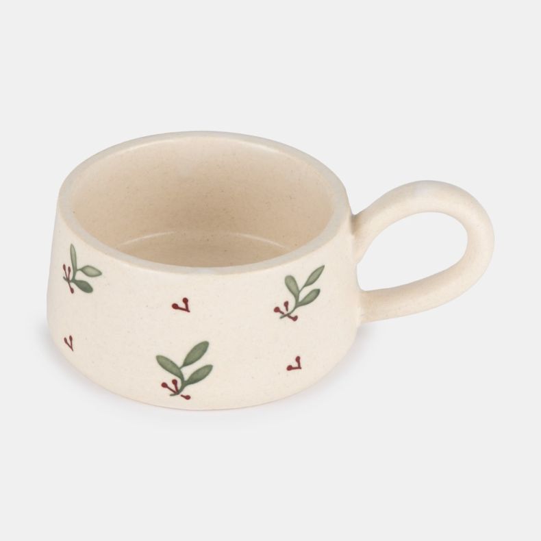 Hand-painted ceramic tealight holder with a rounded loop handle, featuring a cream glazed finish decorated with scattered green leaves and red berry sprigs, shown from an angled view to highlight the botanical design and candle holder shape.