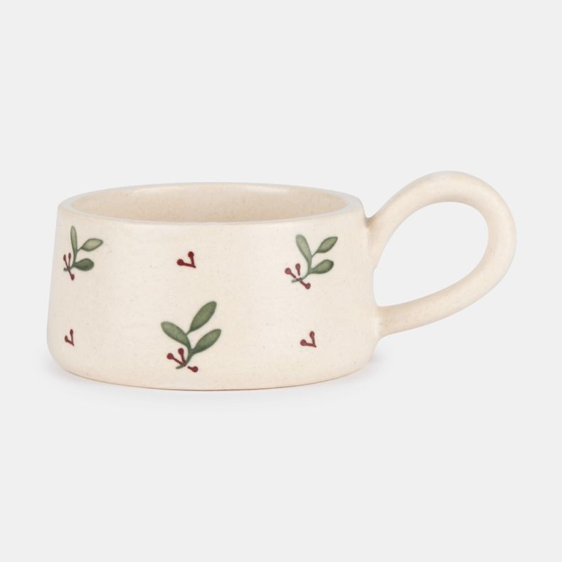 Hand-painted ceramic tealight holder with a rounded loop handle, featuring a cream glazed finish decorated with green leaves and red berry sprigs, shown in side profile to showcase the festive botanical pattern and handcrafted design.