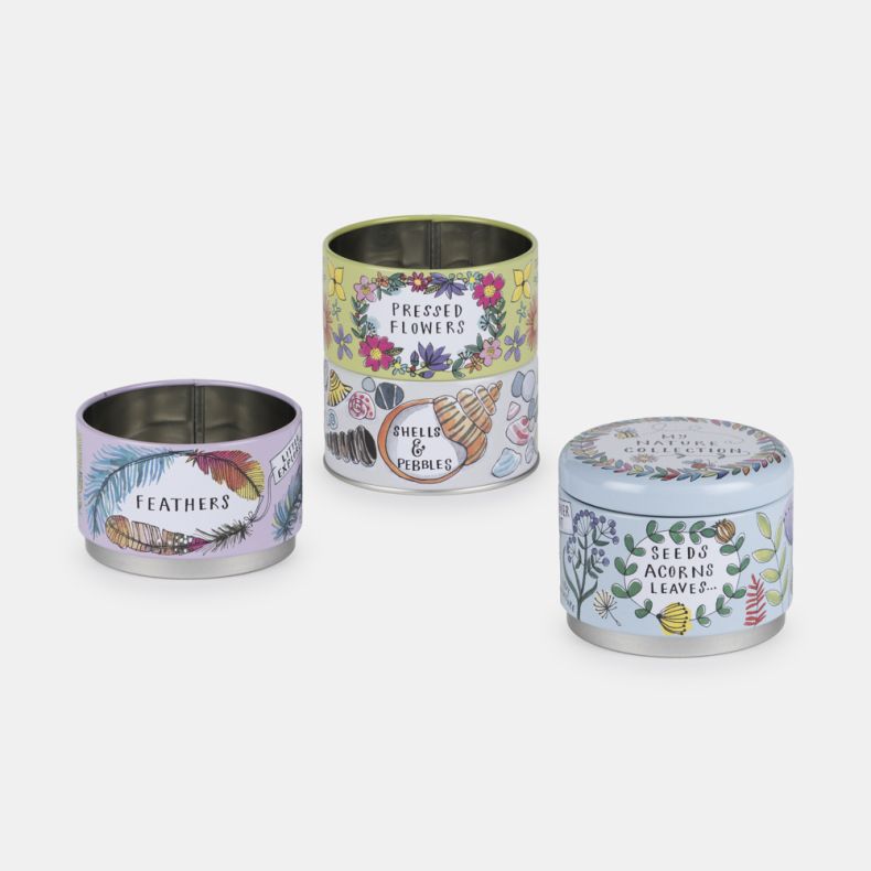 Three separated sections of a nature-themed stackable storage tin collection, decorated with colourful illustrations and labels for “Seeds, Acorns & Leaves,” “Feathers,” “Pressed Flowers,” and “Shells & Pebbles.” The open metal containers are shown agains