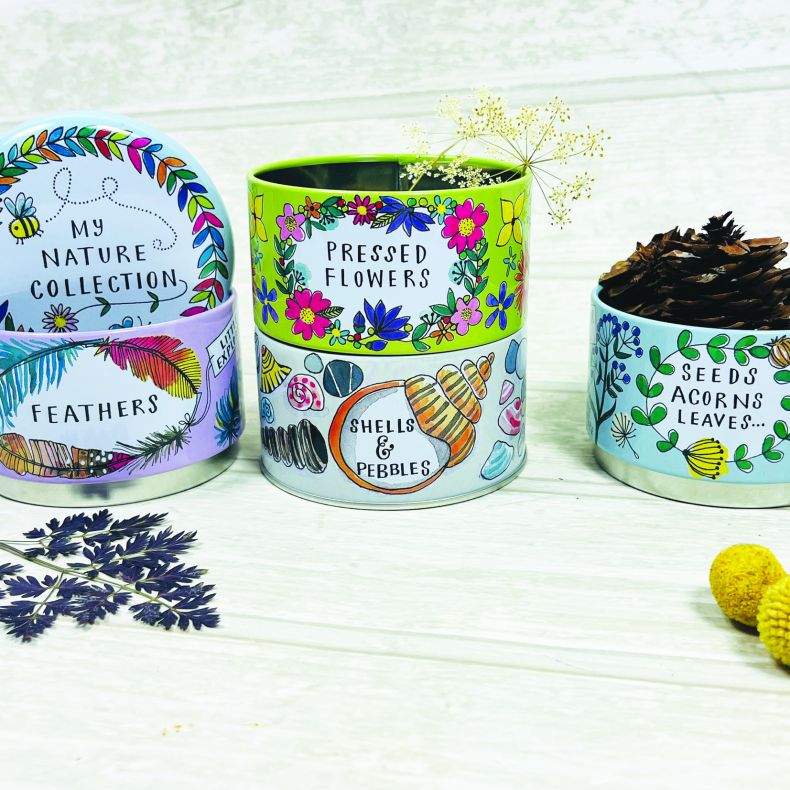 Nature-themed stackable storage tins displayed open and filled with collected treasures. The colourful illustrated containers are labelled “Seeds, Acorns & Leaves,” “Feathers,” “Pressed Flowers,” and “Shells & Pebbles,” with pinecones, dried flowers, and 