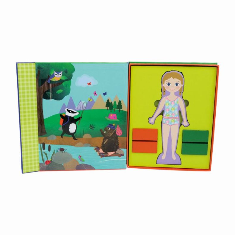 Open magnetic dress-up activity book showing a woodland scene with a badger, bear on a raft, owl in a tree, pond, and mountains on the left page. On the right page, a character figure stands on a yellow background with magnetic outfit pieces stored in red