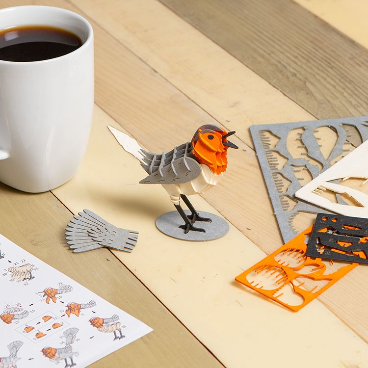 Partially assembled Kikkerland Robin 3D Puzzle displayed on a wooden tabletop alongside instruction sheets and unused puzzle pieces. The robin model features layered grey, white, and orange sections, illustrating the easy-to-build, no-glue assembly proces