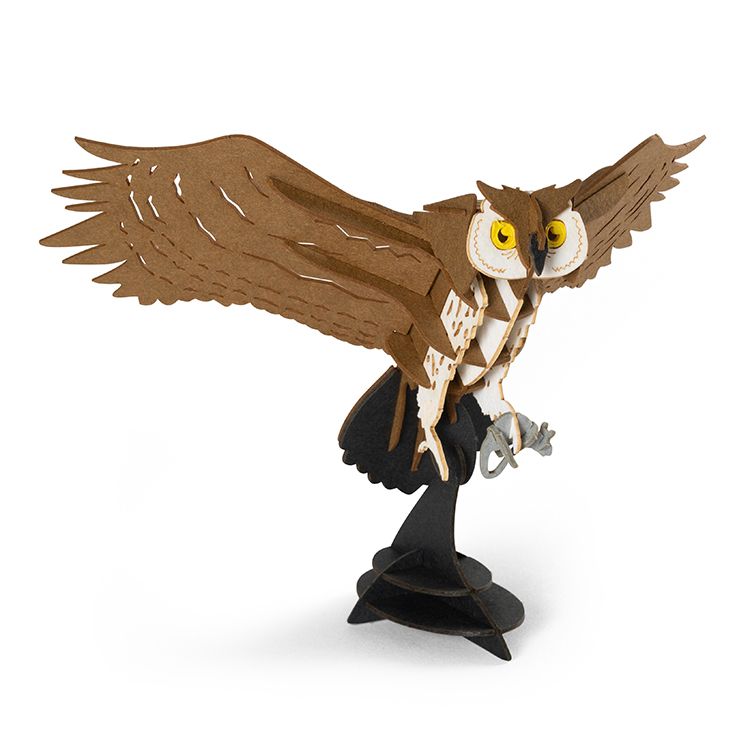 Assembled Kikkerland Owl 3D Puzzle model with wide outstretched wings, bright yellow eyes, and layered brown and cream puzzle pieces. The owl is mounted on a black display stand and shown against a plain light background.