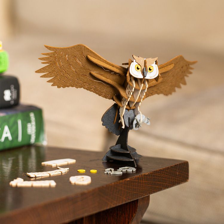 Assembled Kikkerland Owl 3D Puzzle displayed on a tabletop, featuring layered brown wings, cream body details, and bright yellow eyes. Loose puzzle pieces are scattered nearby, showcasing the build process and finished decorative owl model.