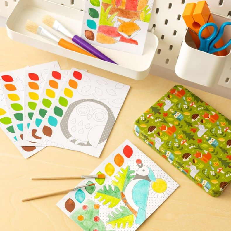 Petit Collage Woodland Paint by Numbers set displayed on a desk, featuring numbered colouring cards, paint palette sheets, two paintbrushes, completed woodland animal artwork, and a decorative storage tin with woodland illustrations.