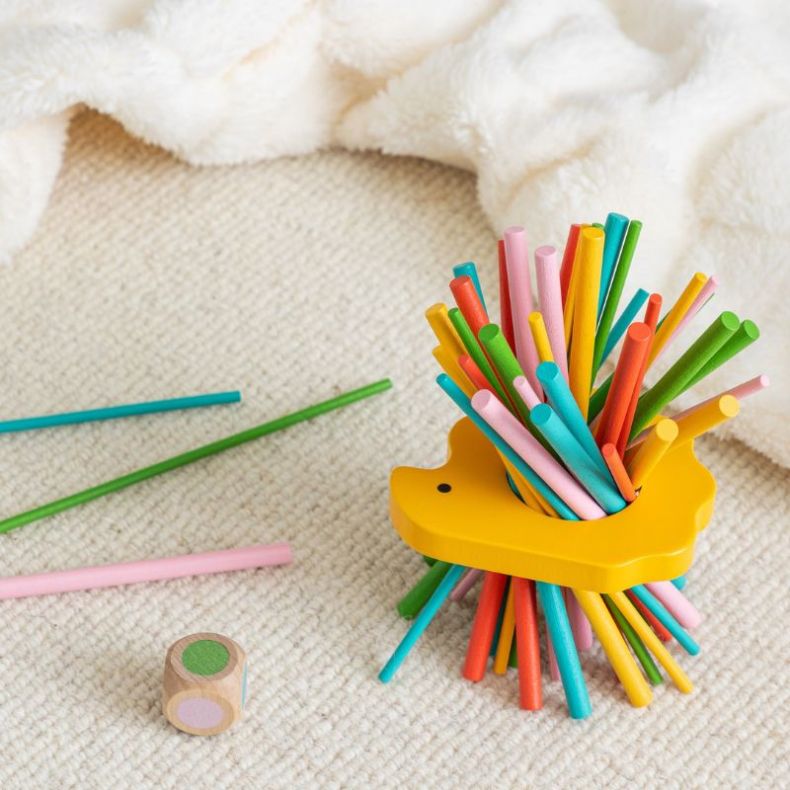 Petit Collage Hedgehog Prickly Sticks game in use, featuring a wooden hedgehog threaded with colourful sticks and a colour-matching die on a soft surface, with additional sticks placed nearby during play.