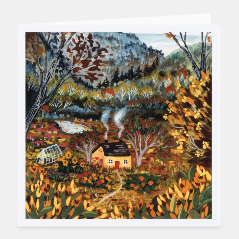 Cosy Cottage notecard featuring a colourful countryside illustration of a small cottage with a red door and smoking chimney, surrounded by flowering gardens, trees and rolling hills in autumnal shades of gold, green, orange and red, displayed with a white