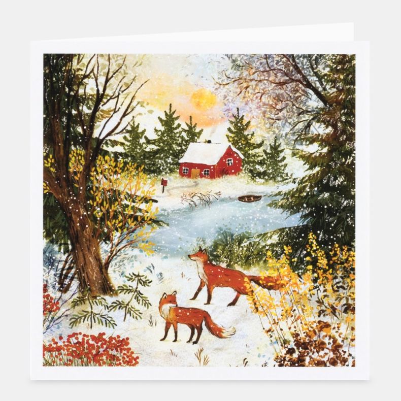 Cosy Cottage notecard featuring a winter countryside illustration of two foxes standing in the snow beside a frozen pond, with a red cottage nestled among evergreen trees in the background, framed by frosted woodland and golden winter sunlight, displayed 