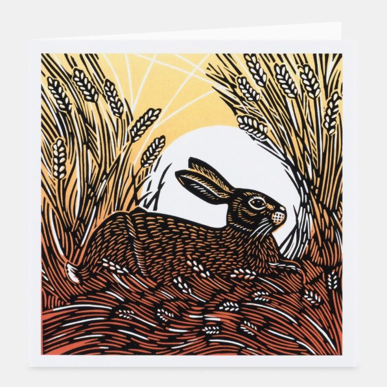 Countryside Encounters notecard featuring a linocut-style illustration of a brown hare resting among golden wheat stems, framed by textured grasses and seed heads with a pale full moon in the background, rendered in shades of gold, cream, brown and black,