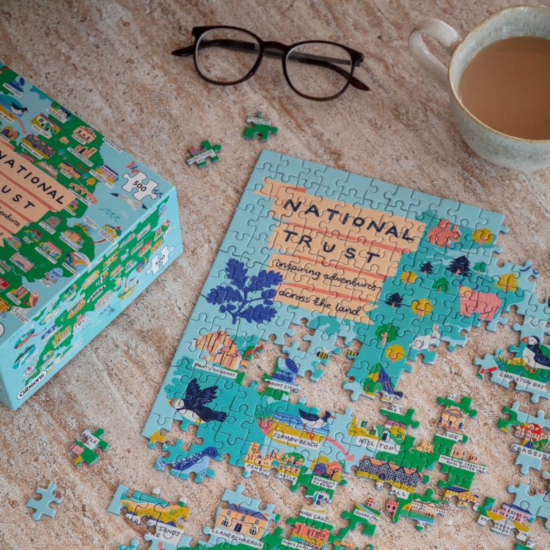National Trust 500-piece illustrated jigsaw puzzle partially assembled on a tabletop, featuring colourful UK landmarks, wildlife and attractions, with puzzle box, glasses and a cup of tea alongside the puzzle pieces