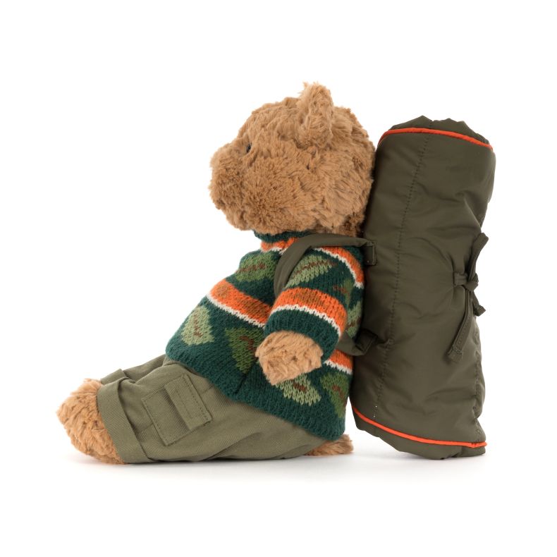 Side view of a Jellycat teddy bear plush with soft beige fur, wearing a green knitted sweater with orange and white stripes and khaki trousers. The bear carries a large olive-green backpack with orange trim and sits against a plain light background.