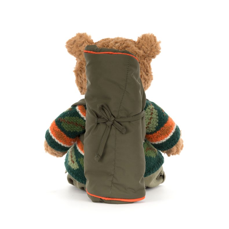 Rear view of a Jellycat teddy bear plush with soft beige fur, wearing a green camouflage-style knitted sweater and khaki trousers. A large olive-green backpack with orange trim is strapped to the bear’s back, shown against a plain light background.