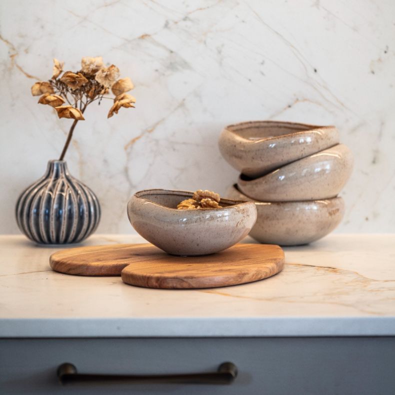 Speckled ceramic serving bowl collection styled on a light marble-effect worktop in a contemporary kitchen setting. In the foreground, a rounded beige ceramic bowl with an organic irregular rim holds walnut halves and is displayed on a pair of overlapping