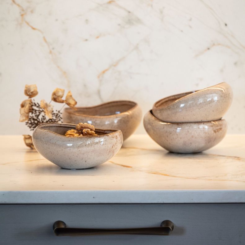 Collection of speckled ceramic serving bowls displayed on a light marble-effect surface in a styled kitchen setting. The bowls feature an organic rounded shape with gently irregular rims, a glossy beige glaze and delicate speckled detailing throughout. On