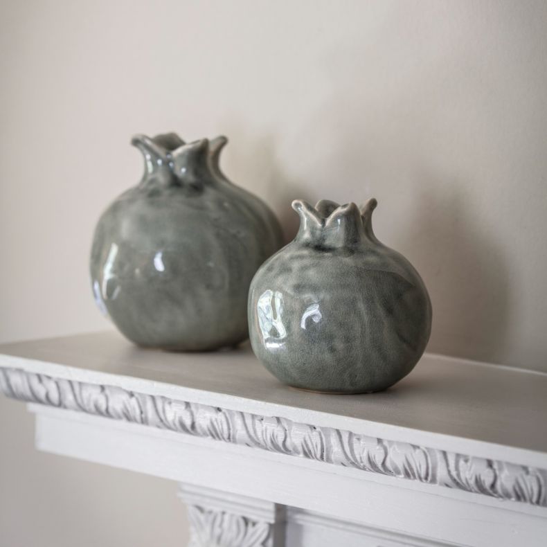 Two decorative ceramic vases styled on a white mantelpiece in a softly lit interior. Each vase features a rounded, pomegranate-inspired silhouette with a distinctive crown-shaped opening and a glossy blue-grey glaze with subtle tonal variations and a deli