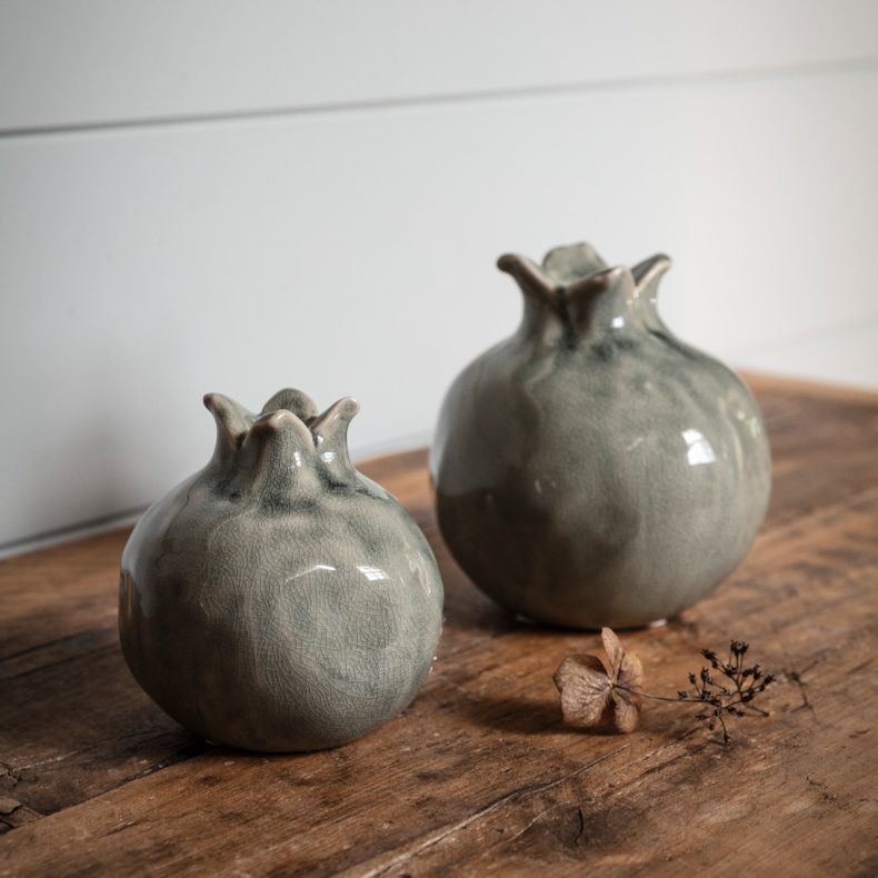 Two decorative ceramic vases with rounded, pomegranate-inspired shapes displayed on a rustic wooden tabletop. Each vase features a distinctive crown-shaped opening and a glossy blue-grey glaze with subtle tonal variations and a visible crackle-effect fini