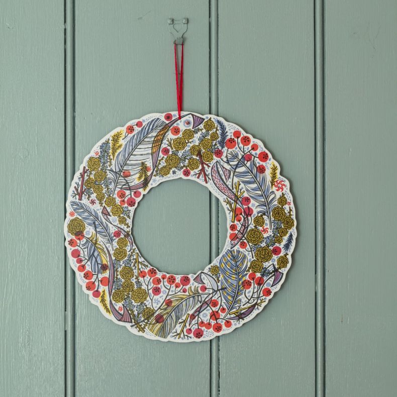 Festive wreath-shaped advent calendar decoration featuring illustrated feathers, berries and winter foliage in red, blue and gold tones, finished with a red hanging ribbon and displayed against a painted wooden backdrop.