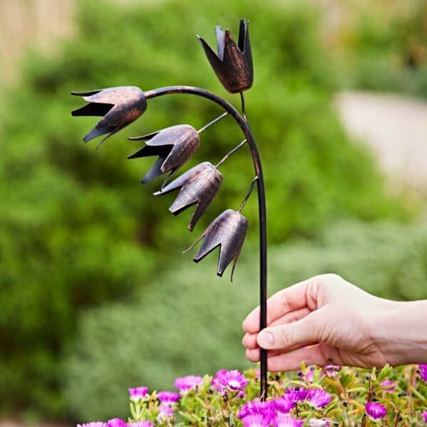 Metal Bluebell Garden Plant Stake being placed into a flower bed, featuring five handcrafted bell-shaped blooms with a rustic bronze-effect finish on curved stems, shown alongside a hand for scale in an outdoor garden setting.