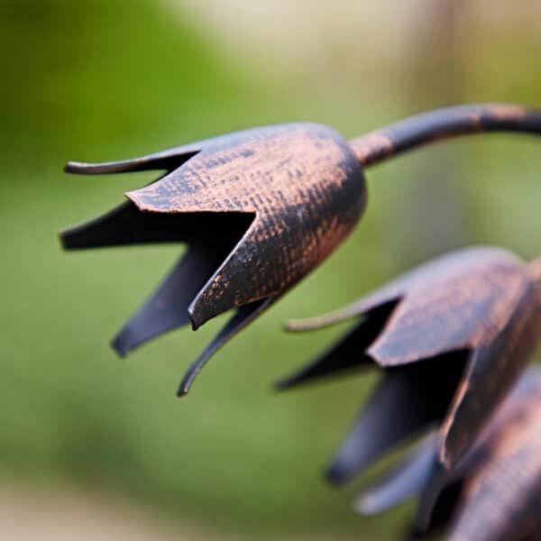Close-up detail of the Metal Bluebell Garden Plant Stake, showing handcrafted bell-shaped metal flowers with a rustic bronze-effect finish and textured petals on curved stems against a softly blurred garden background.