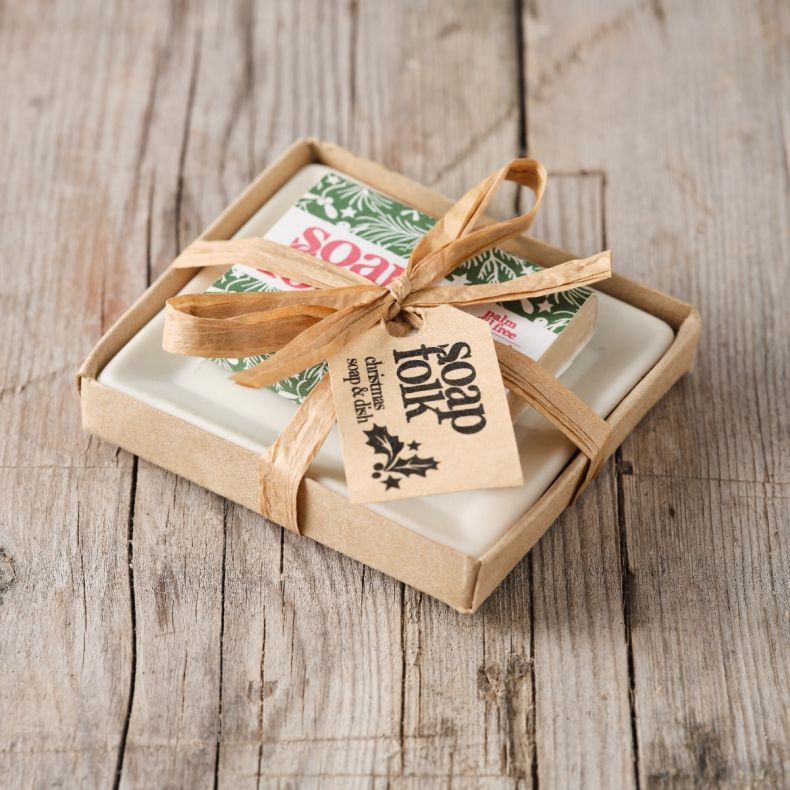 Soap Folk Christmas soap and soap dish gift set displayed on a rustic wooden surface, featuring a cream ceramic soap dish and a Christmas-themed soap bar wrapped in green festive packaging, presented in a kraft gift box and tied with a natural ribbon and 