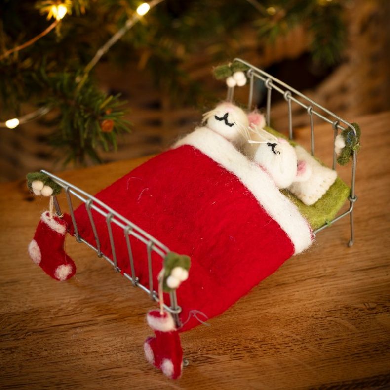 Felt mice Christmas hanging decoration in a miniature metal bed, featuring two white wool-effect mice tucked beneath a red felt blanket, with embroidered closed-eye detailing, pink ears, whisker accents, green felt pillows, festive stocking decorations, a