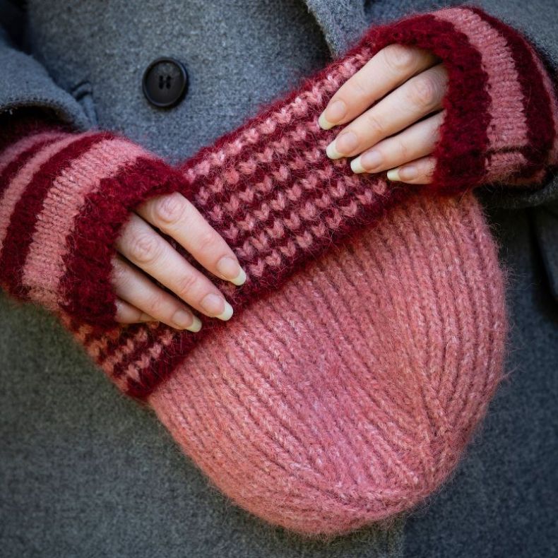 Close up of person holding a pink ribbed knit beanie hat with a chunky textured turn-up brim in contrasting burgundy and pink stripes, featuring a soft fluffy yarn with matching hand warmers.