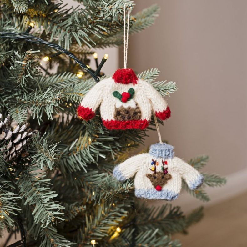 Set of two knitted Christmas jumper hanging decorations displayed on a Christmas tree, featuring a festive Christmas pudding design with holly leaves and red berries, and a reindeer design with a red nose and multicoloured lights on the antlers, suspended