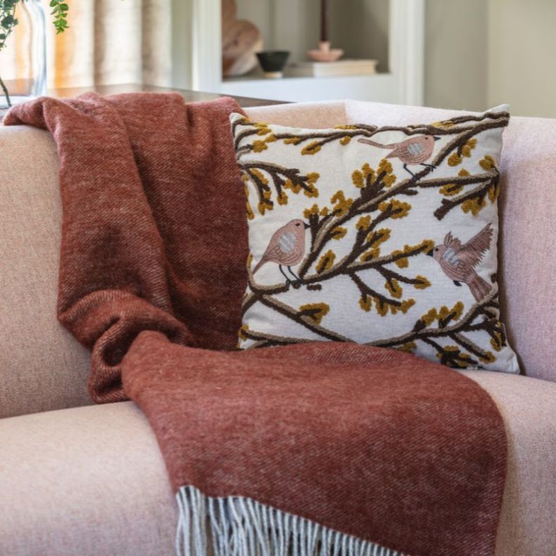 Rust-coloured wool throw draped over a pale pink sofa, styled beside an embroidered cushion featuring birds perched on branching foliage in soft pink, brown and mustard tones, displayed in a cosy living room setting.