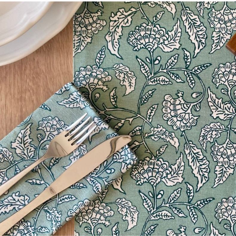 Green floral patterned cotton napkin and table runner set featuring an illustrated botanical design in sage green, styled with silver cutlery on a wooden tabletop to showcase the detailed print and fabric texture.