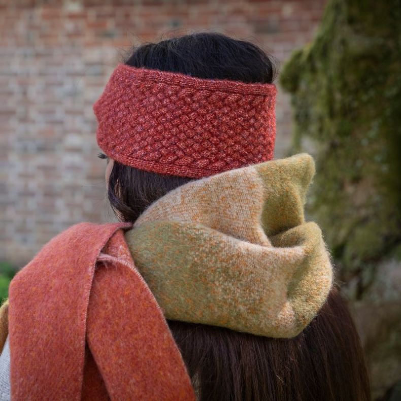 Rust textured knitted headband in a chunky weave, shown worn outdoors from the rear with a soft green and rust colour-block scarf to highlight the coordinated winter accessories and textured knit design.