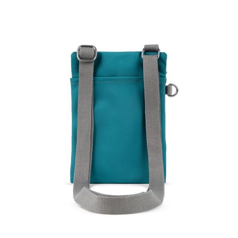 Teal crossbody pouch shown in angled view, featuring a grey shoulder strap, front zip pocket, and ROKA London label.