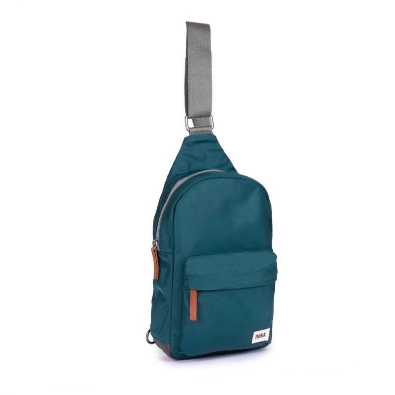 Teal sling backpack shown in angled view, featuring a wide grey shoulder strap, front zip pocket, brown zip pull tabs, and a side attachment ring.