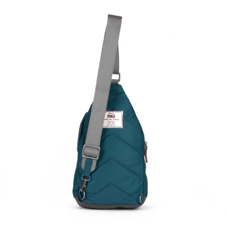 Teal sling backpack shown from the rear, featuring a wide grey adjustable shoulder strap, quilted back panel, and metal attachment rings.