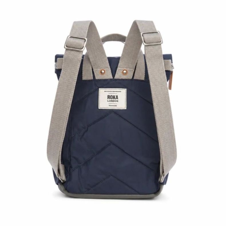 Roka London backpack shown from the back in navy blue, featuring adjustable light grey shoulder straps, quilted back panel and top carry handle. Lightweight design with a rear Roka London logo patch and durable water-resistant finish.
