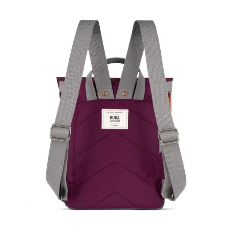 rear view of a burgundy red Roka London backpack featuring adjustable grey shoulder straps, padded back panel with stitched chevron detailing, top carry handles, and a woven Roka London logo label. The roll-top backpack is shown against a plain light back