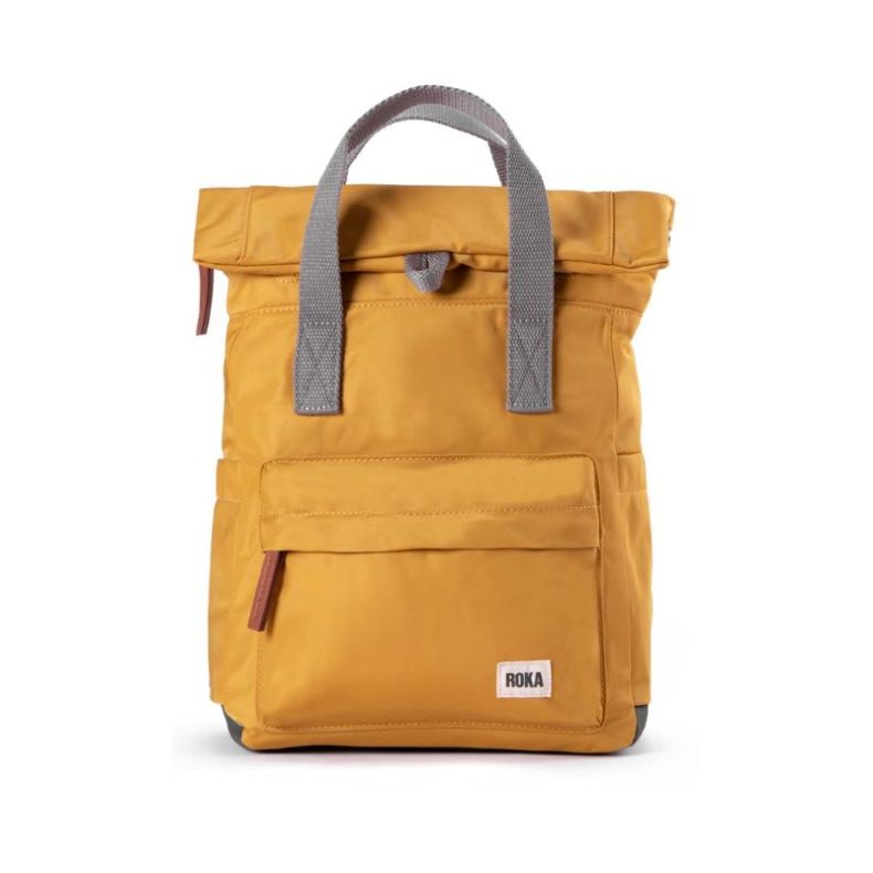 A stylish mustard-yellow backpack with a modern roll-top design, featuring grey fabric handles and a front zippered pocket. The backpack has a minimalist aesthetic with a small "ROKA" brand label on the lower front.