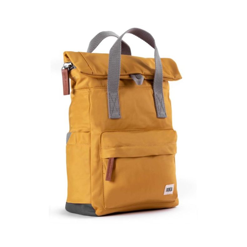 A mustard-yellow backpack with a modern, minimalist design, photographed from a three-quarter angle. The bag features a roll-top closure secured with a grey fabric loop, two sturdy grey handles, and a front zippered pocket with a leather pull tab.