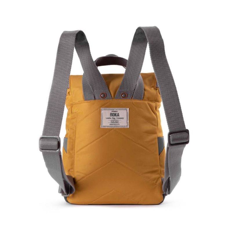 A mustard-yellow backpack viewed from the back, featuring adjustable grey fabric straps and a reinforced top handle. The back panel has a quilted chevron pattern for added comfort. A rectangular "ROKA" brand label is stitched onto the upper section