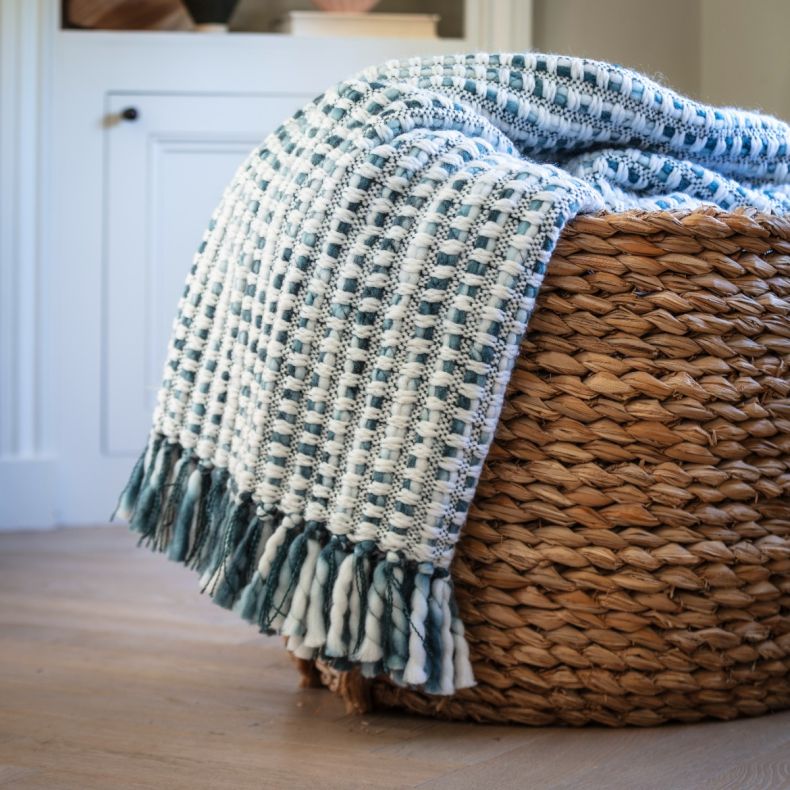Blue and cream woven throw blanket with chunky textured weave and tasselled fringe, draped over a woven storage basket to showcase the cosy fabric, contrasting colours and decorative tassel detailing.