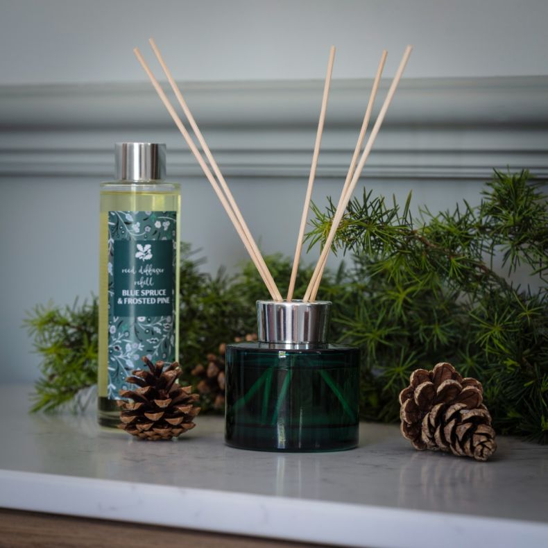 Blue Spruce & Frosted Pine reed diffuser in a dark green glass vessel with natural reed sticks, displayed alongside a matching refill bottle, pine cones and evergreen foliage to showcase the festive home fragrance collection.