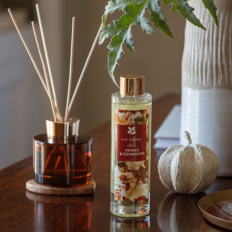 Orange & Cedarwood reed diffuser refill in a clear glass bottle with autumn leaf design label, displayed alongside a matching amber glass reed diffuser, decorative pumpkin and vase to showcase the seasonal home fragrance collection.