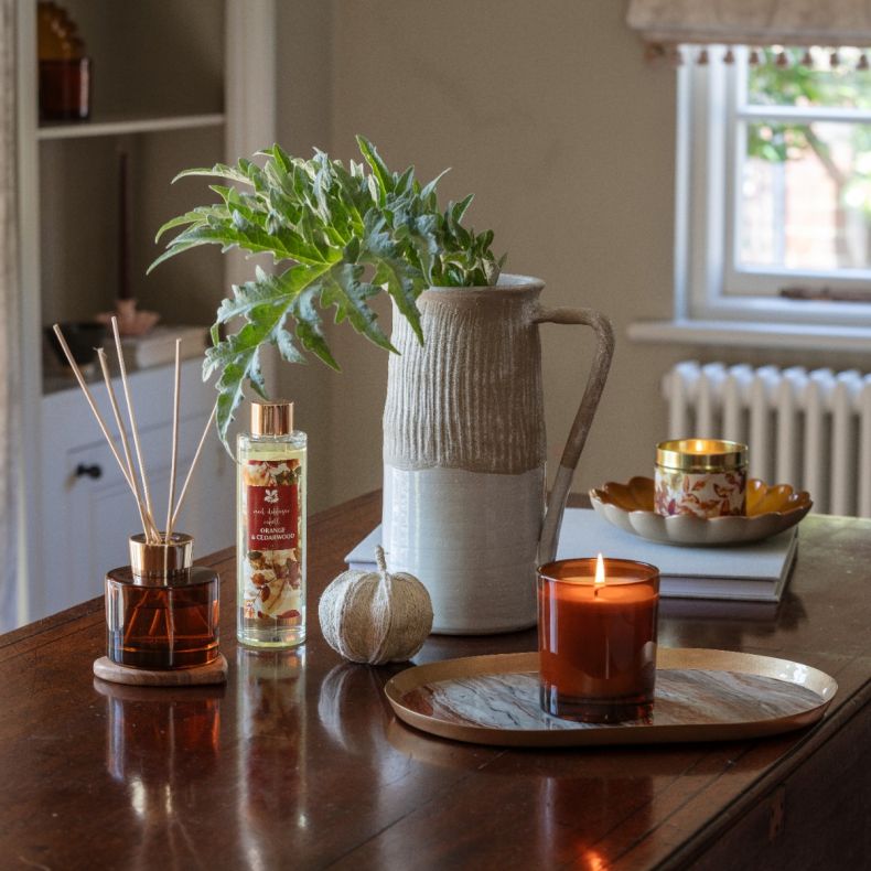 Orange & Cedarwood home fragrance collection displayed on a wooden table, featuring an amber glass reed diffuser, matching refill bottle and lit candle alongside a ceramic jug vase with foliage, decorative pumpkin and autumn-inspired styling.