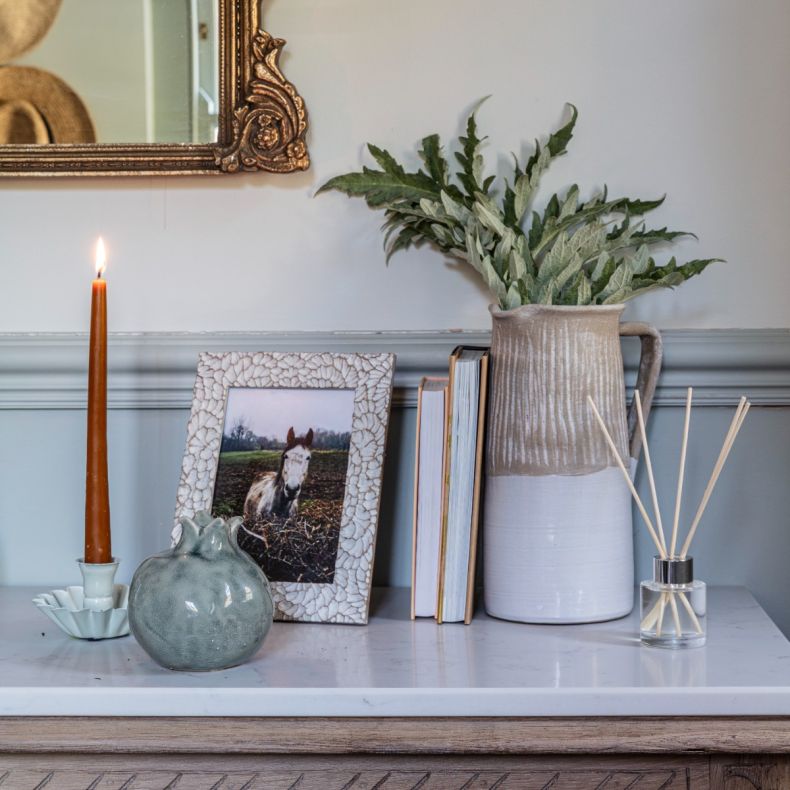 Small reed diffuser with natural reeds in a clear glass bottle, styled on a mantelpiece alongside books, a textured ceramic jug vase with foliage, a decorative pomegranate ornament, framed horse photograph and lit taper candle in a rustic home interior.
