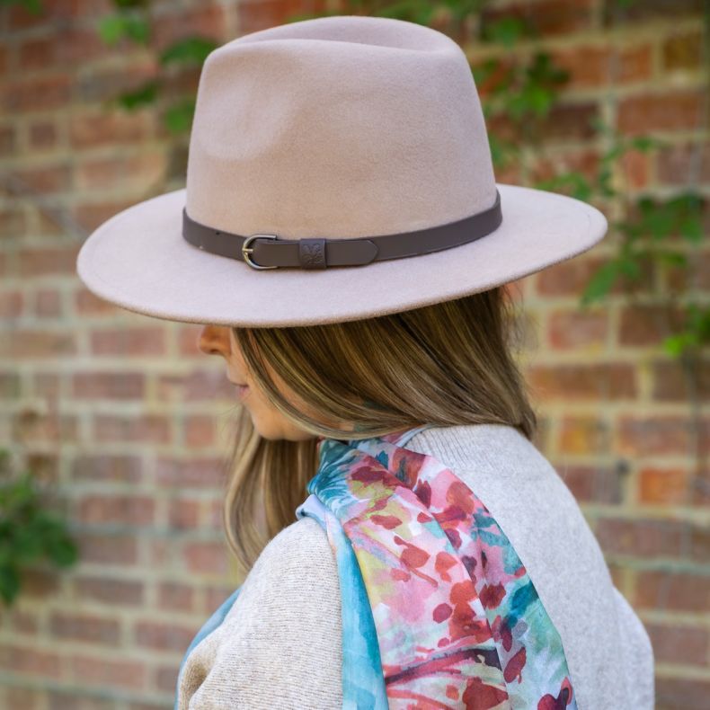 Stone beige felt fedora hat with a wide brim and brown buckle band, shown worn outdoors in side profile view with a colourful floral print scarf to highlight the classic shape and coordinating autumn accessory styling.